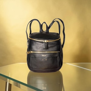 May include: A black leather backpack with a pebbled texture, featuring gold-tone metal zippers, a top handle, and adjustable shoulder straps. The front displays the brand name PERLINA in gold lettering above a small pocket with two gold studs.