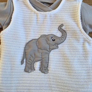 Op de afbeelding: Een witte mouwloze romper van geribbelde stof met een grijze olifantapplicatie op de voorkant. Het kledingstuk heeft een ronde halslijn met overlappende schouderdetails en wordt gedragen over een grijs shirt met lange mouwen.
