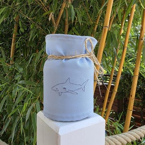 May include: A light blue fabric cover for a jar or container, featuring a dark grey embroidered shark silhouette on the front. The top of the fabric is gathered and secured with a piece of natural tan twine tied in a simple bow.
