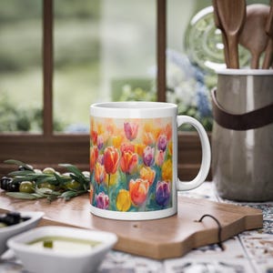 May include: White ceramic mug with a watercolour painting of tulips in red, orange, yellow, and purple. The mug sits on a wooden board, with olives and olive oil in the foreground. The mug is approximately 9 cm tall.