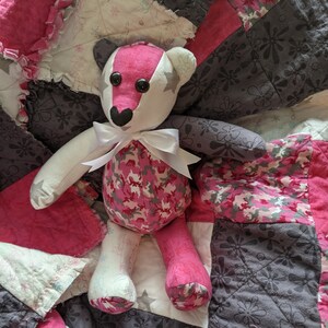 May include: A pink and white camouflage teddy bear with black eyes and a white bow, sitting on a pink and grey patchwork quilt.