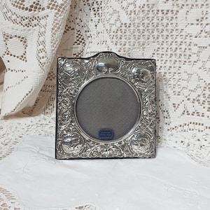 May include: Square silver-toned picture frame with an ornate floral and scroll design. The frame has a circular opening with a grey backing. A small blue label is visible at the bottom. The frame is displayed on a white surface.