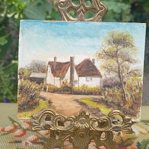 May include: A small painting on canvas depicting a white cottage with a thatched roof, surrounded by trees and a dirt path. The painting is framed and displayed on a gold easel.