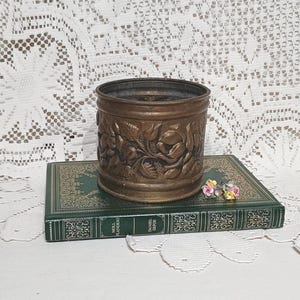 May include: A vintage bronze planter with floral relief sits on a green book. The cylindrical planter features a detailed pattern of roses and leaves. Small, colourful flowers rest on the book's cover.