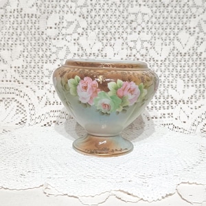 May include: A vintage porcelain vase with a pedestal base. The vase has a light blue and brown background, decorated with pink and green roses. The rim is trimmed with gold detailing.