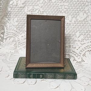 May include: A bronze-coloured rectangular picture frame with a dark interior sits on a green book with gold lettering. The frame has a detailed, ornate border. The book has decorative gold patterns.