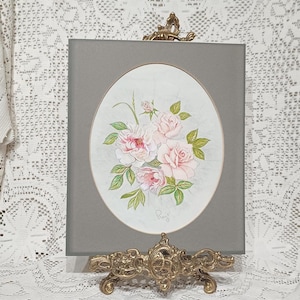 May include: A framed oval print of pink roses and green leaves, displayed on a gold-toned easel. The artwork has a grey border and is set against a white lace tablecloth. The overall style is vintage, with a focus on floral design.