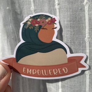 Pu&ograve; includere: Un adesivo con una donna che indossa un velo turchese e una corona di fiori. La donna guarda di lato e l'adesivo ha la parola "EMPOWERED" scritta su uno stendardo rosso.