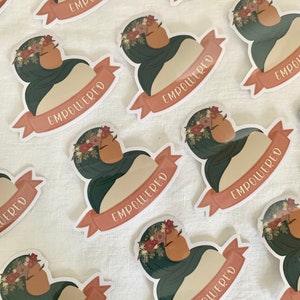 Pu&ograve; includere: Un set di adesivi con una donna che indossa un velo verde e una corona di fiori. Gli adesivi hanno uno striscione color pesca con la parola "EMPOWERED" scritta in bianco.
