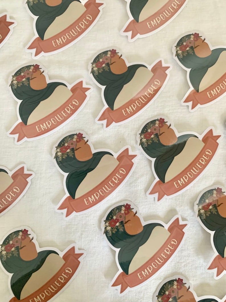 Pu&ograve; includere: Un set di adesivi con una donna che indossa un velo verde e una corona di fiori. Gli adesivi hanno uno striscione color pesca con la parola "EMPOWERED" scritta in bianco.
