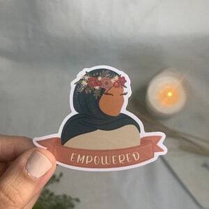 Pu&ograve; includere: Un adesivo con una donna che indossa un velo blu e una corona di fiori. L'adesivo ha uno striscione che recita "EMPOWERED".