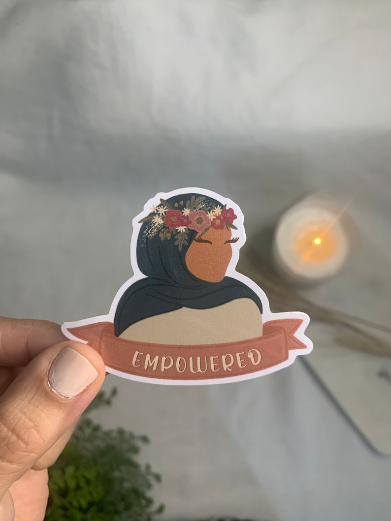 Pu&ograve; includere: Un adesivo con una donna che indossa un velo blu e una corona di fiori. L'adesivo ha uno striscione che recita "EMPOWERED".