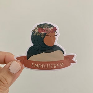 Pu&ograve; includere: Un adesivo con una donna che indossa un velo verde e una corona di fiori. L'adesivo ha uno striscione rosso con la parola "EMPOWERED" stampata su di esso.