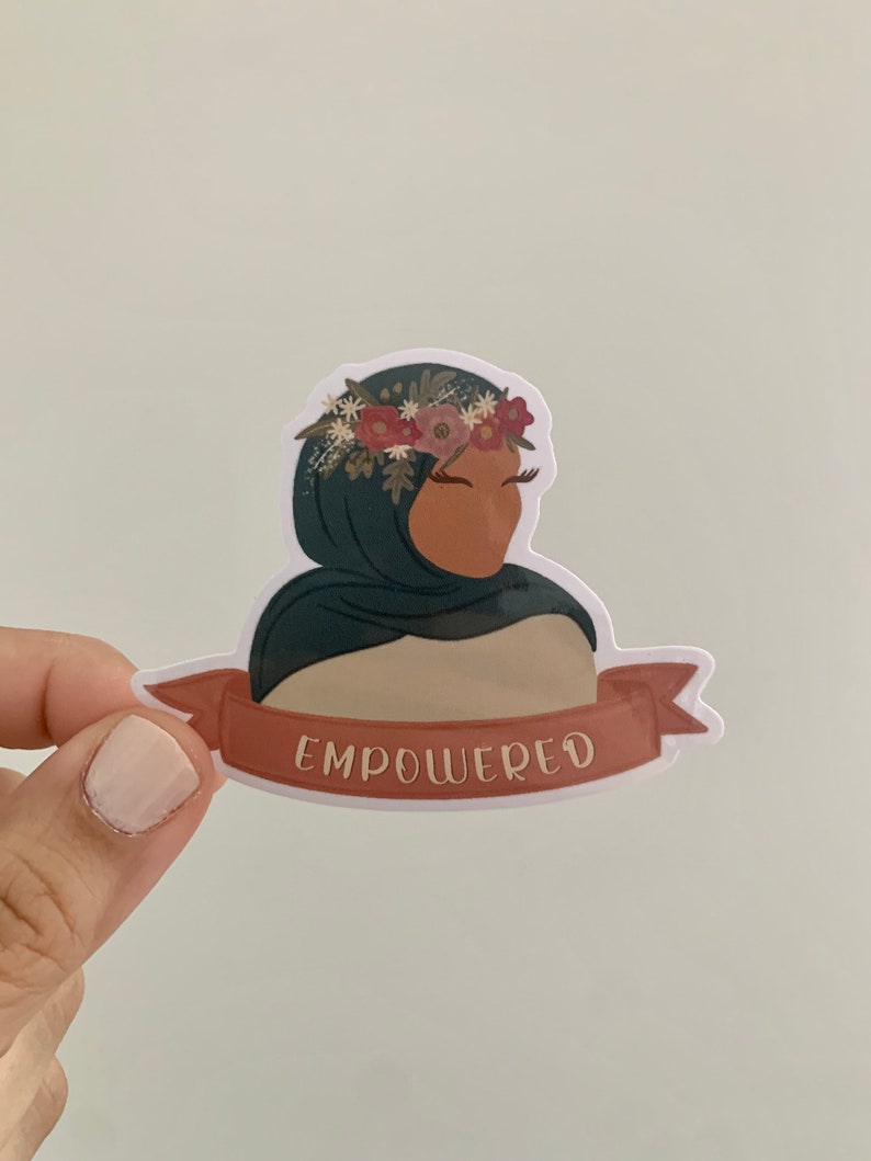 Pu&ograve; includere: Un adesivo con una donna che indossa un velo verde e una corona di fiori. L'adesivo ha uno striscione rosso con la parola "EMPOWERED" stampata su di esso.