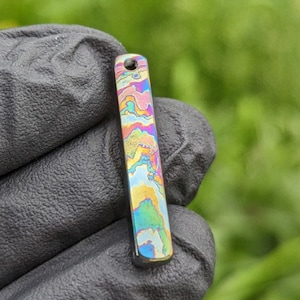 Könnte beinhalten: Ein bunter, schillernder Metallanhänger in Form eines Rechtecks mit abgerundeten Kanten. Der Anhänger hat ein wirbelndes Muster aus Blau, Grün, Gelb und Rosa.