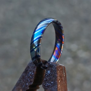 Könnte beinhalten: Nahaufnahme eines blauen und violetten gemusterten Metallrings, der in einer Metallzange gehalten wird. Der Ring ist glänzend und hat eine glatte Oberfläche.