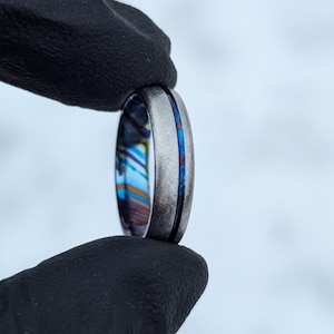 Könnte beinhalten: Ein silberfarbener Ring mit einem lebendigen, mehrfarbigen Innenraum und einem dünnen blauen Streifen in der Mitte. Der Ring wird zwischen zwei schwarzen behandschuhten Fingern gehalten, was sein kompliziertes Design zeigt. Der Ring hat Blau-, Orange- und Gelbtöne.