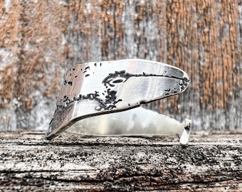 May include: A handcrafted silver ring with a unique, textured design. The ring features an abstract shape with dark accents, set against a rustic wooden background. The ring's design is artistic and eye-catching.