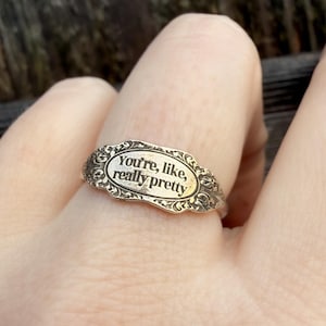 Könnte beinhalten: Silberfarbener Ring mit ovaler Oberfläche. Auf der Oberfläche des Rings steht in einer Serifenschrift "You're, like, really pretty". Der Ring hat ein verziertes, florales Design um das Oval.