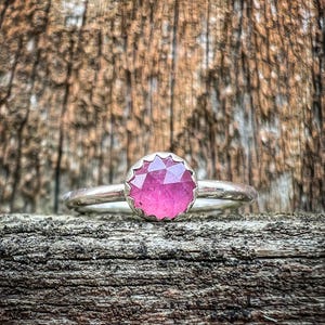Puede incluir: Un anillo de plata con una piedra preciosa rosa engastada en un engaste facetado. El anillo está apoyado en un trozo de madera desgastada.