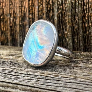 Pode incluir: Um anel de prata com uma grande pedra da lua oval. A pedra da lua exibe um jogo de cores iridescente, com tons de azul, verde e amarelo. O anel está sobre uma superfície de madeira, realçando a beleza natural da gema.