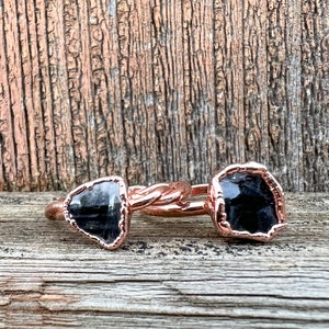 May include: Two black gemstone rings with rose gold bands. The rings are twisted together and are sitting on a wooden surface.