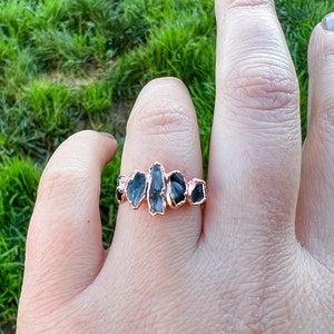 May include: A rose gold ring featuring a cluster of five black gemstones. The gemstones are irregularly shaped and have a raw, natural appearance.