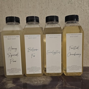May include: Four clear plastic bottles of "THE NINTH FLAME MOP SOAP" with black lids. Each bottle has a white label with a different scent: Honey Spiced Pear, Balsam Fir, Eucalyptus, and Frosted Cranberry. The bottles contain a light yellow liquid.
