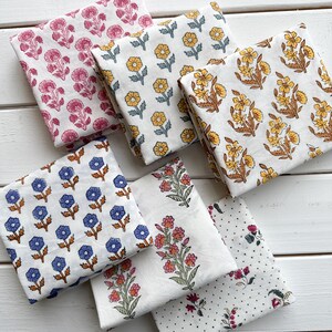 May include: Six folded squares of white fabric featuring various floral block prints. The patterns include pink carnations, yellow daisies, yellow and brown wildflowers, blue flowers with brown stems, pink and green floral sprigs, and small black dots.