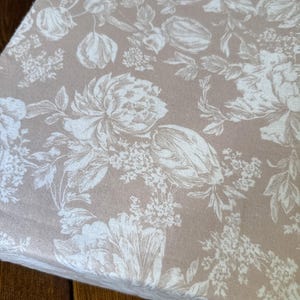 May include: A folded piece of fabric featuring a light beige background with a white floral pattern. The design consists of detailed illustrations of various flowers, leaves, and stems distributed across the entire surface of the textile material.