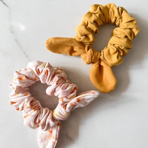 Puede incluir: Dos coleteros con lazos. Uno es amarillo mostaza, el otro es rosa con un estampado floral naranja. Los coleteros están hechos de tela suave y se exhiben sobre una superficie de mármol blanco.