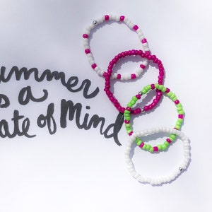 May include: Four colorful beaded bracelets stacked on top of each other. The bracelets are white, pink, and green. The text "Summer is a state of mind" is written in black ink on a white background.