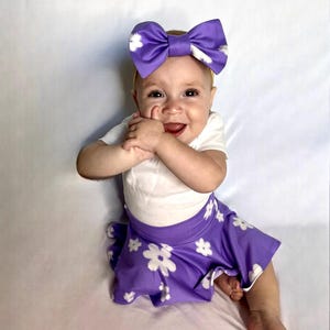 May include: A baby girl wearing a white shirt and a purple skirt with white flowers. She is wearing a purple bow headband with white polka dots.