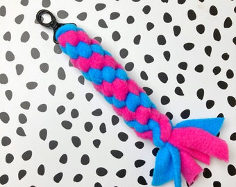 May include: A braided dog toy in bright pink and blue fleece. The toy has a black clip at the top and a knotted end with blue and pink streamers. The background is white with black polka dots.