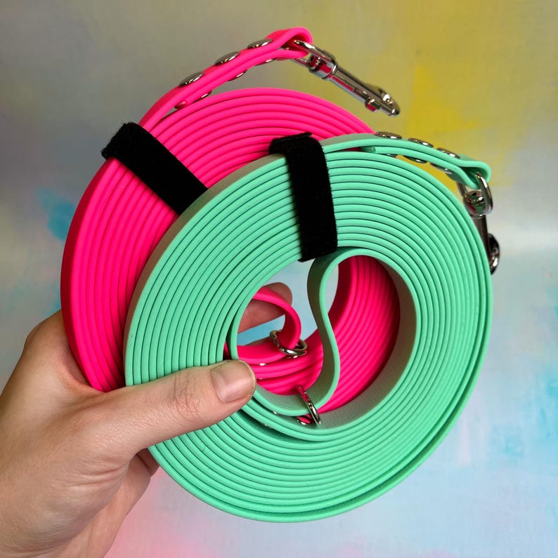 May include: Two dog leashes, one pink and one green, are rolled up together. The leashes are made of a thick, durable material and have a black strap.