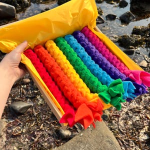 May include: A cardboard box lined with bright yellow tissue paper containing six long, braided fleece ropes in a rainbow gradient of red, orange, yellow, green, blue, and purple. Each rope features knotted ends made of soft fabric.