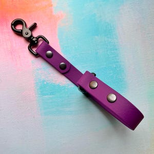 May include: A purple strap with a matte finish featuring a black metal swivel lobster clasp at one end. The strap is secured with four circular silver-toned metal rivets arranged in pairs along the length of the purple material.