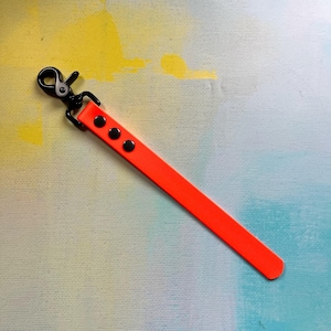 May include: A bright neon orange wrist strap featuring a matte black metal swivel lobster clasp. The strap is secured with three black circular rivets arranged in a vertical line near the top, providing a clean and functional design.