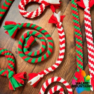 May include: A collection of braided rope dog toys made from red, green, and white fabric strips. The toys feature various knotted and twisted designs with frayed ends. Some are coiled, while others are straight, showcasing a woven texture.