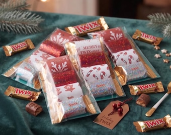 May include: Three clear plastic gift bags containing Twix candy bars and decorative inserts with red, white, and glittery patterns. The center bag features the text MERRY TWIX-mas. Loose Twix bars, a small gift tag labeled Holiday TWIX, and a gold spoon are present.