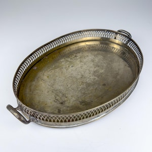 Könnte beinhalten: Ein ovales Serviertablett aus Metall mit angelaufener, messingfarbener Oberfläche. Das Tablett verfügt über eine dekorative, durchbrochene Galerieumrandung und zwei kleine, geschwungene Griffe an den gegenüberliegenden Enden.