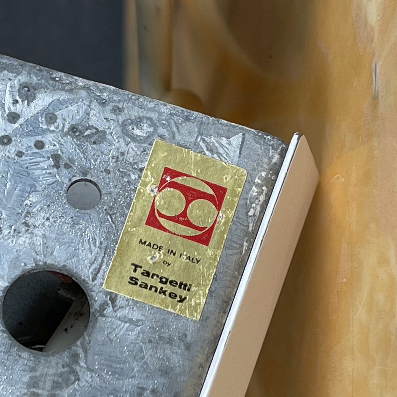 Pode incluir: Um r&oacute;tulo dourado com um logotipo vermelho e o texto "Made in Italy by Targetti Sankey" em uma superf&iacute;cie de metal prateado.