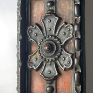 Pu&ograve; includere: Primo piano di un pannello decorativo in metallo con un ornato motivo floreale grigio scuro. Il pannello ha uno sfondo color rame ed &egrave; incorniciato da un bordo scuro e strutturato. L'elemento floreale &egrave; centrato e dettagliato con piccoli punti in rilievo.