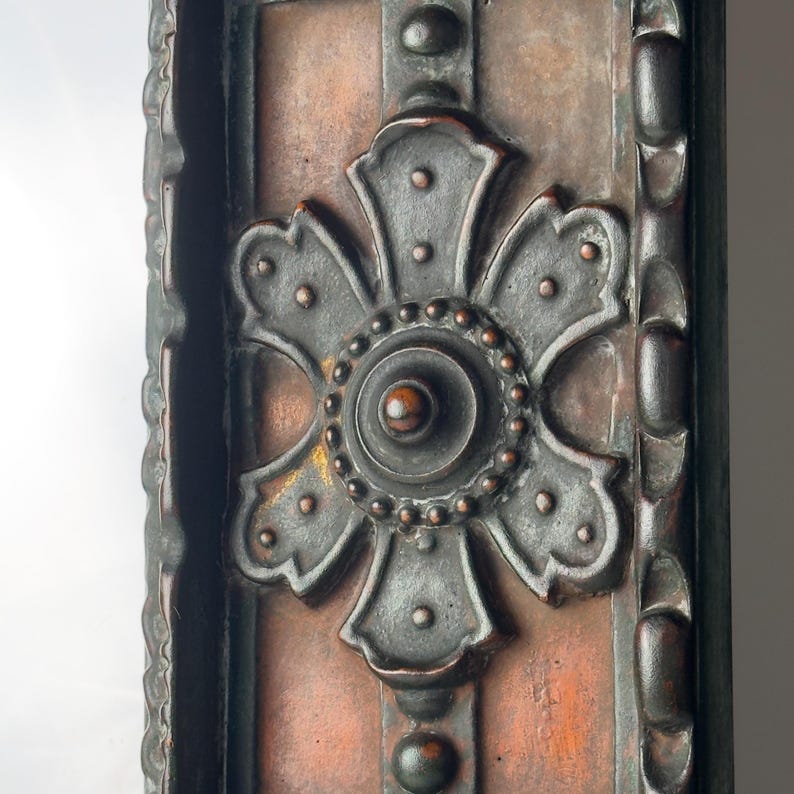Pu&ograve; includere: Primo piano di un pannello decorativo in metallo con un ornato motivo floreale grigio scuro. Il pannello ha uno sfondo color rame ed &egrave; incorniciato da un bordo scuro e strutturato. L'elemento floreale &egrave; centrato e dettagliato con piccoli punti in rilievo.