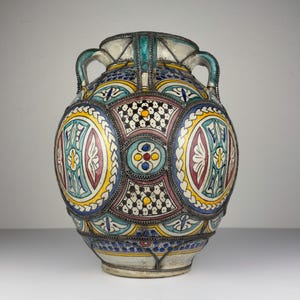 Pode incluir: Um vaso de cerâmica com duas alças, apresentando um padrão geométrico complexo em amarelo, azul-petróleo, azul e branco. A superfície é dividida em painéis circulares e retangulares delineados por filigrana de metal escuro e pequenos motivos florais.