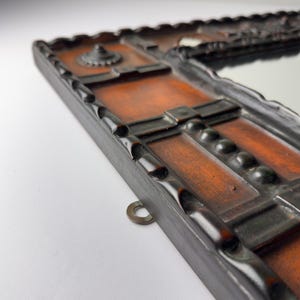 Pu&ograve; includere: Primo piano di una cornice rettangolare in legno per specchio con finitura marrone scuro. La cornice presenta un design decorativo intagliato con uno specchio centrale. Un anello di metallo &egrave; attaccato alla cornice per appenderla. La cornice ha un motivo geometrico dettagliato.