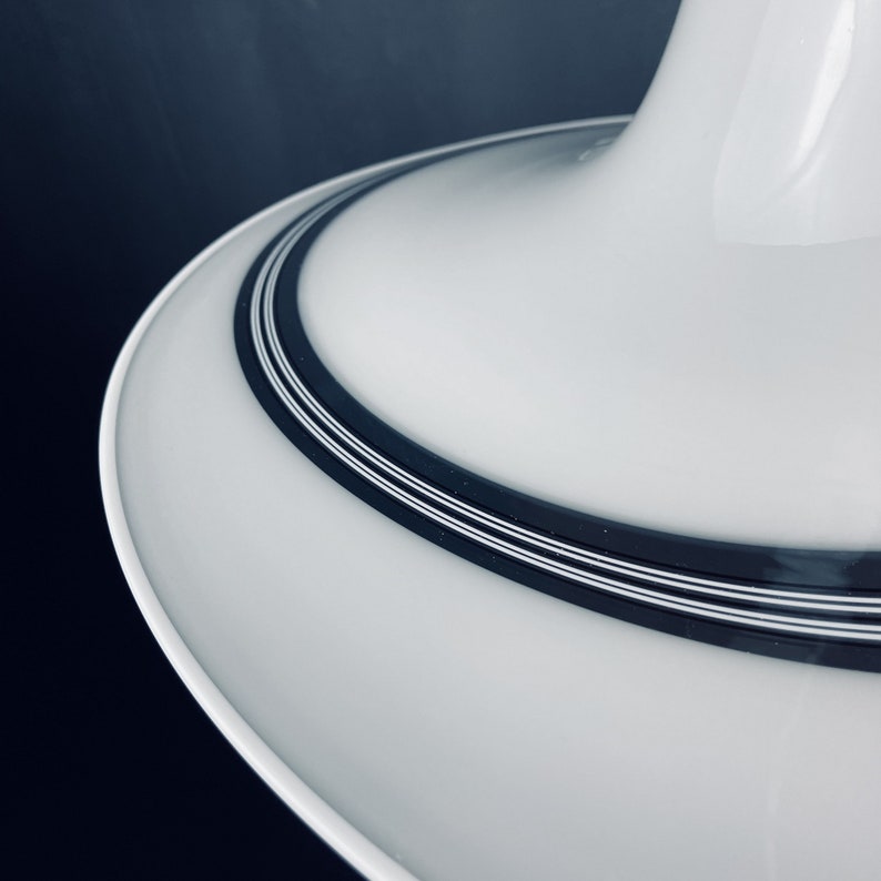 May include: Close-up of a white ceiling light fitting with a black and white striped trim. The light fitting has a wide, flat, circular base and a smaller, round, cone-shaped top.