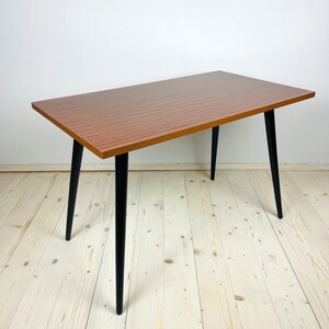 Peut inclure: Une table rectangulaire en bois avec un plateau en stratifi&eacute; brun et des pieds effil&eacute;s noirs. La table est pos&eacute;e sur un sol en bois clair.