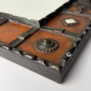 Pu&ograve; includere: Primo piano di una cornice decorativa per specchio con un'estetica rustica e antica. La cornice presenta una superficie strutturata marrone scuro con accenti metallici e un bordo nero. Il design include un ornamento centrale circolare e un piccolo dettaglio a forma di diamante.