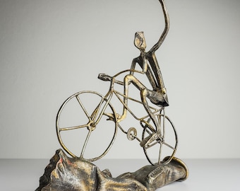 Può includere: Scultura in metallo di un ciclista su bicicletta, con un braccio alzato. La scultura è principalmente dorata e argentata, con una base strutturata. La bicicletta ha ruote grandi e dettagli intricati. Un oggetto decorativo.